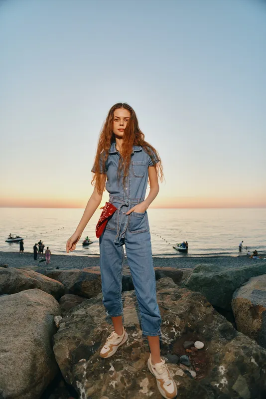 Lifestyle woman in modern boho-western denim street style stands confidently on rocks by the sea at sunset, showcasing casual film color fashion and relaxed summer vibes.