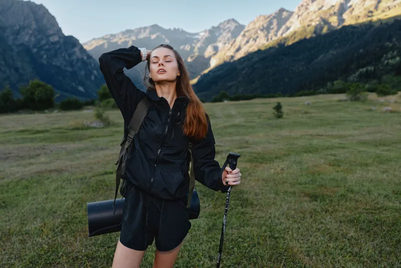 An adventurous woman stands in a scenic mountain meadow, holding a trekking pole and wearing a backpack. She looks confident, energized, and ready for outdoor exploration.
