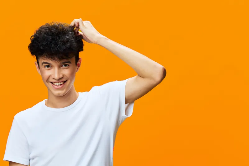 smiling man in white t-shirt scratching head with curly hair on bright orange background, cheerful young adult, casual style, isolated studio portrait