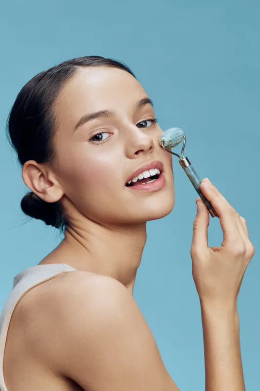 Young woman with a joyful expression using a facial roller against a pastel blue background, promoting skincare and beauty routines for healthy skin