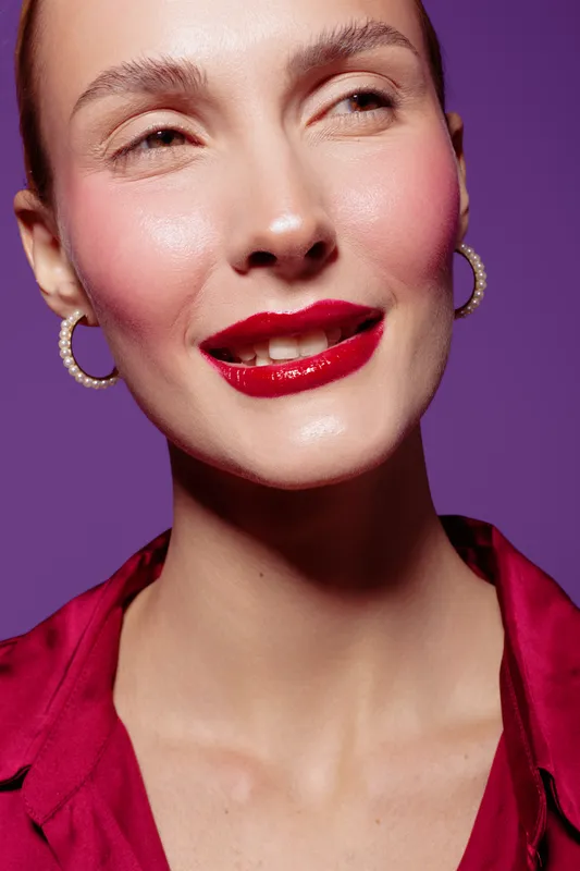 Close-up portrait of a smiling woman with glossy red lips and hoop earrings wearing a red blouse. Studio shot with purple background. Concept of beauty, fashion, and makeup.