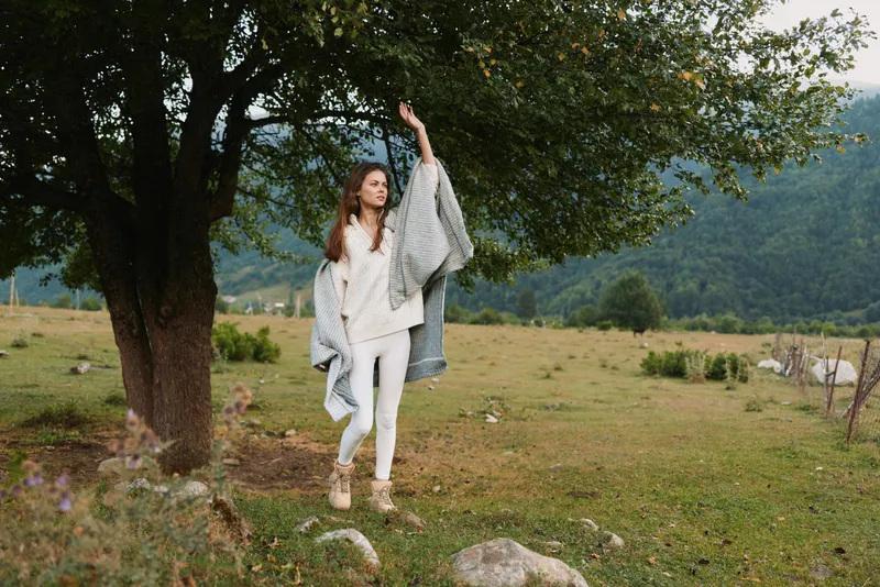 Young woman in white walks beside a tree in a sunlit field, lifting a blanket as if dancing, embodying carefree countryside vibes.