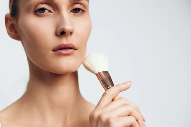 Young woman applies makeup with a brush on her face in a close up studio shot, calm expression, soft lighting highlighting smooth skin and natural tones