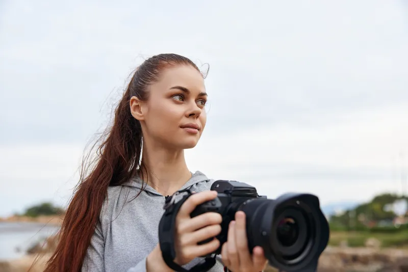 Young woman with long hair holding a camera outdoors, showcasing creativity and passion for photography with a calm expression against a natural background