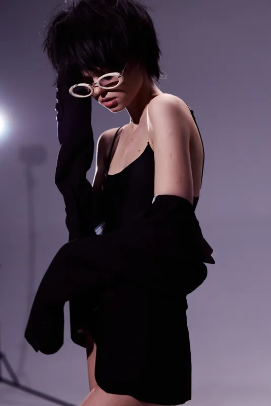Stylish woman in black dress with glasses posing against neutral background, elegant look, modern fashion, studio lighting