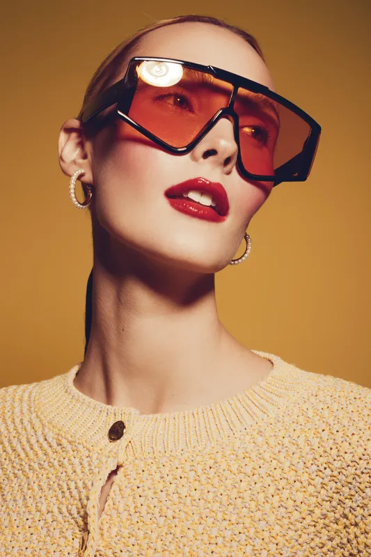 Stylish woman with oversized orange sunglasses and red lipstick poses against yellow background. She wears gold hoop earrings and a textured beige sweater. Fashion and beauty concept.