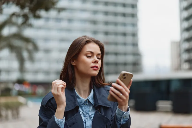 Young woman with long brunette hair wearing a dark coat looks thoughtfully at her smartphone while standing in an urban environment with blurred buildings in the background.