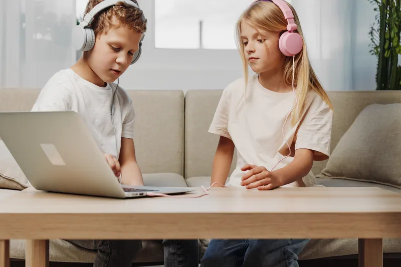 Two children with headphones use laptop at wooden table in cozy living room, focused on online activities and digital entertainment together.