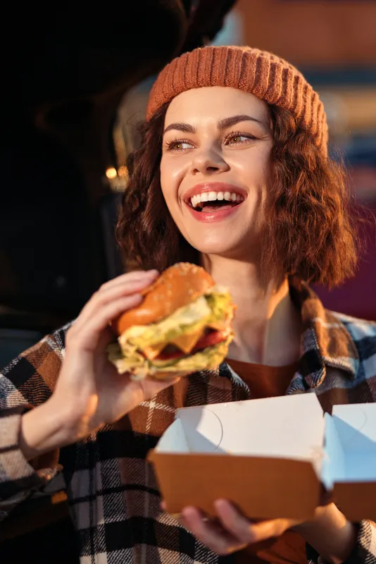 Woman with beanie holding a burger and takeaway box, candid smile and golden hour glow capturing authenticity, candid lifestyle, mindful living and emotional storytelling in urban scene.