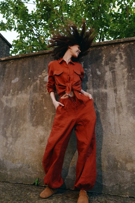 A woman in a bold red jumpsuit stands against a concrete wall, hair tossed by wind, striking a dynamic pose. A confident, energetic street style moment outdoors