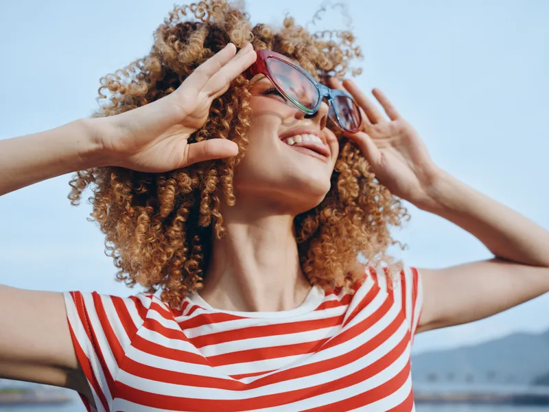 Joyful woman with curly hair wears a red and white striped shirt and sunglasses, enjoying a bright sunny day outdoors, smiling confidently toward the clear sky.