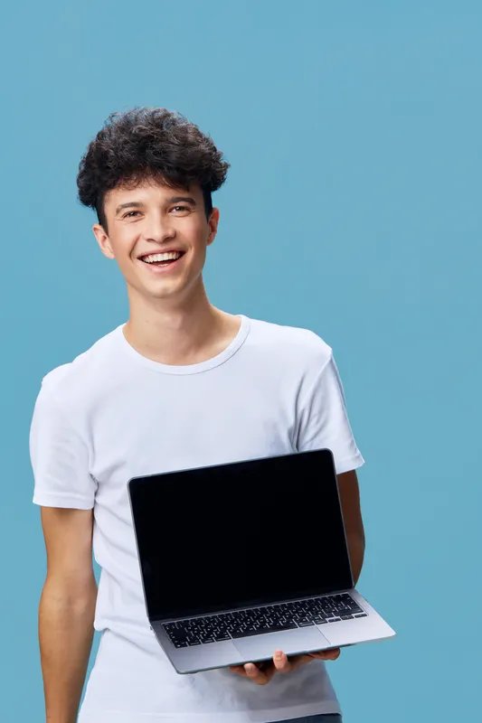 smiling man holding laptop wearing white t-shirt with curly hair against blue background, modern technology and casual style, positive expression