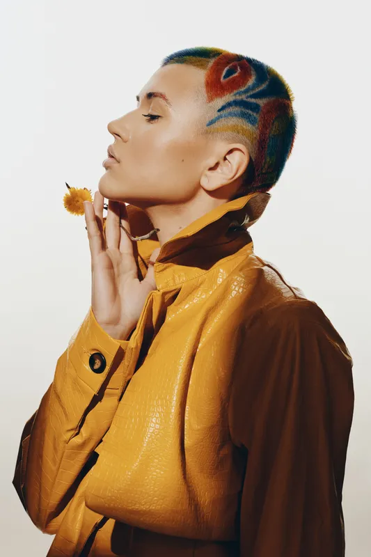 Artistic woman with vibrant colorful shaved hair and yellow textured jacket posing with closed eyes, holding a dried flower near her face in a minimalistic studio setting.
