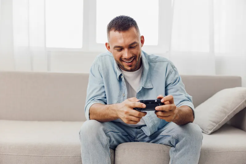 Smiling man sitting on a sofa, joyfully playing video games. Relaxed atmosphere with soft natural light streaming through the window, enhancing the cozy setting.