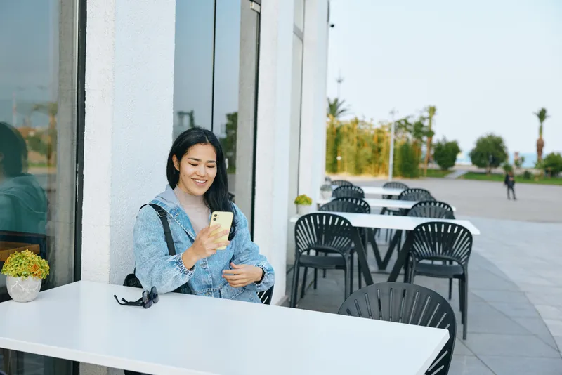 Smartphone led moment as a woman in a blue jacket sits at an outdoor cafe table, smiling at her screen, with patio seating and glass storefronts along a bright sidewalk.
