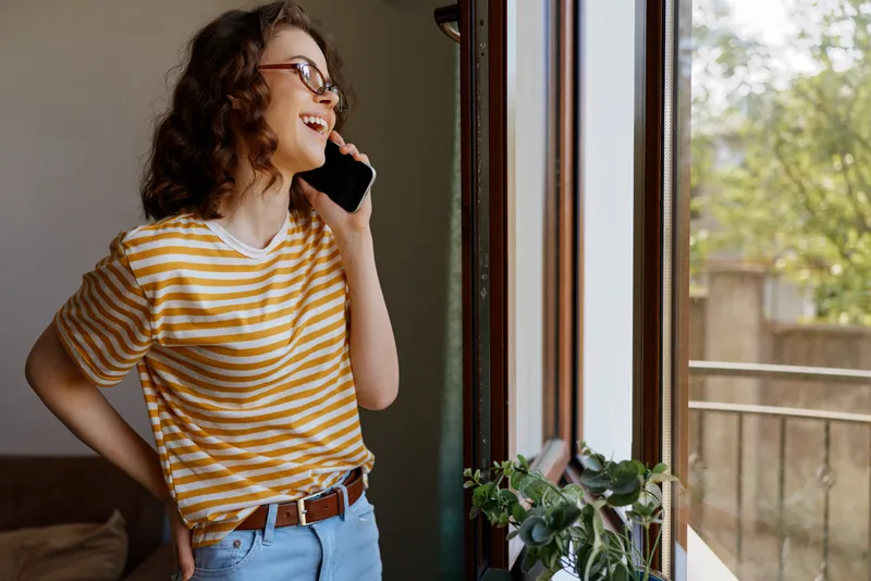 Cheerful woman indoors, talking on phone near sunlit window, striped shirt, jeans, casual home setting, relaxed posture, warm light, natural colors, plant accents, candid lifestyle moment