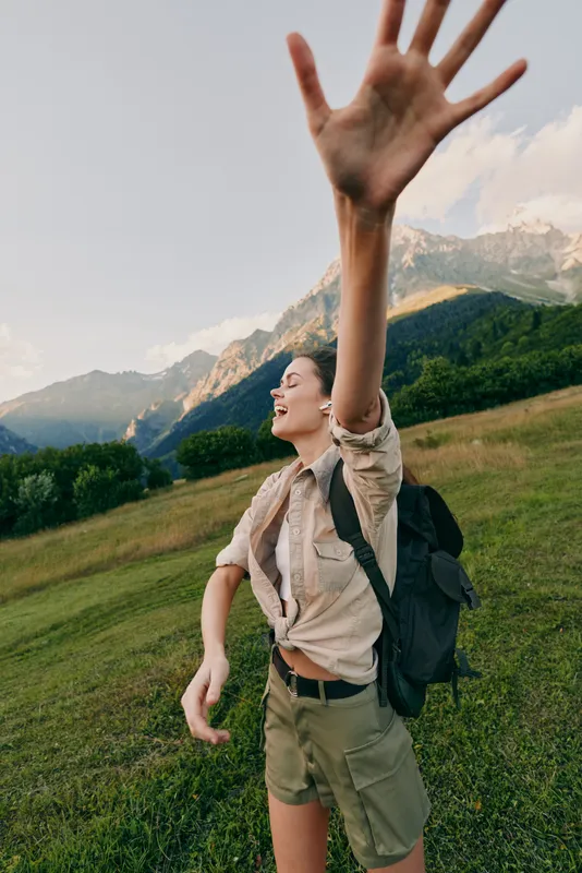Cheerful woman outdoors, hiking with backpack, raising hand in a joyful moment, sunlit meadow with mountain backdrop, natural colors, energetic and carefree lifestyle amid green hills and breeze