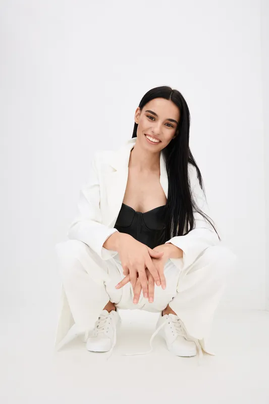 smiling woman with long black hair wearing white suit and black top sitting casually against white background in modern studio setting