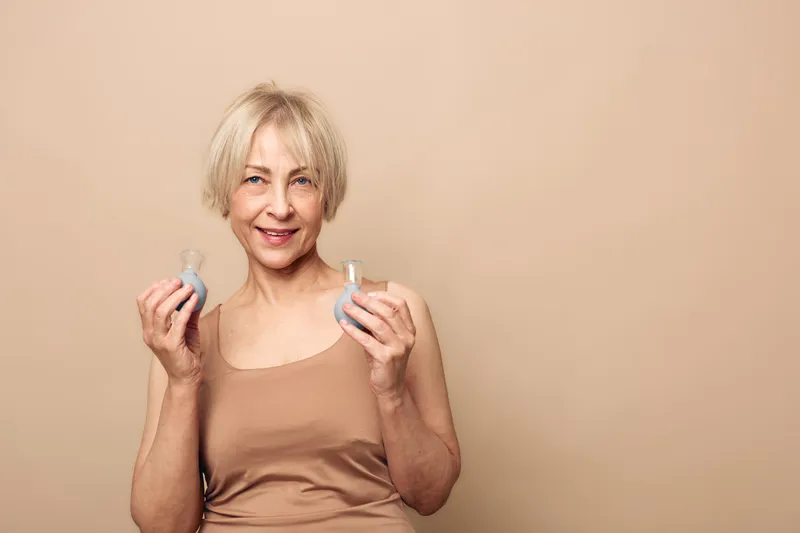 Smiling mature woman with short blonde hair holds two small cosmetic containers in hands, wearing beige top against plain beige background. Concept of skincare, beauty, and self-care routine.
