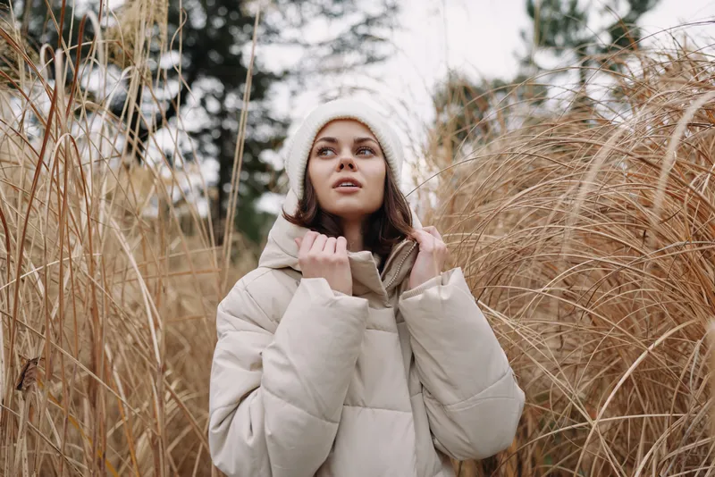 Woman in a warm coat stands among tall dried grass outdoors, gazing upward with a calm expression, capturing a winter mood, natural setting, street style meets countryside serenity