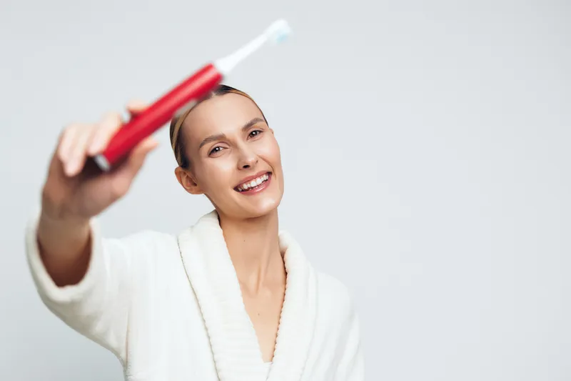 Smiling woman in white bathrobe holding red toothpaste tube, fresh clean face, bright background, personal care, morning routine, dental hygiene, healthy lifestyle concept.