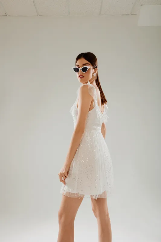 Young woman in a white lace dress poses confidently wearing stylish sunglasses. Studio shot on a neutral background. Concept of woman beauty fashion and elegance.