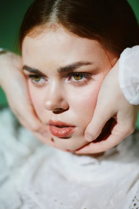 Captivating portrait of a young woman with expressive eyes, wearing a delicate white blouse, showcasing emotion and vulnerability against a soft green background