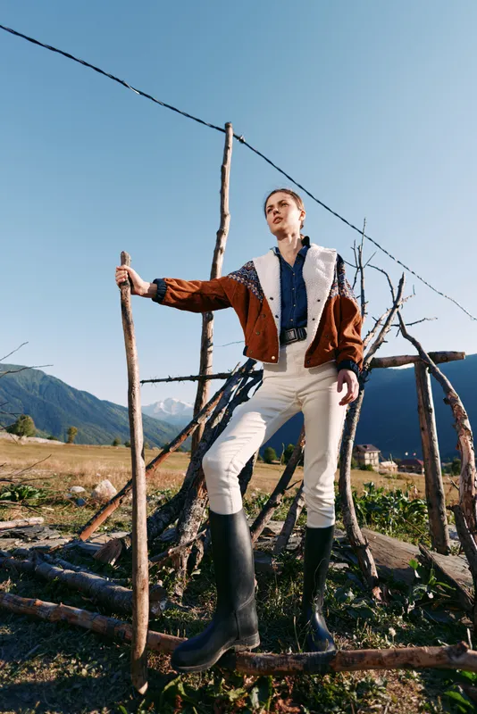 woman rural mountains walking boots stick fence, young female standing in countryside with hiking stick, wearing rubber boots and warm jacket, portrait in open landscape under clear sky