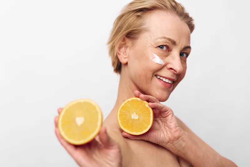 Smiling mature woman with short blonde hair holding two halves of an orange close to her face with cream applied on cheek, isolated on white background. Concept of skincare and natural beauty.
