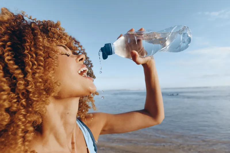 Cheerful woman outdoors, drinking water from a bottle at the sunny beach, energetic moment, natural light, wind in hair, clear sky, blue sea, relaxed pose and reflections on water