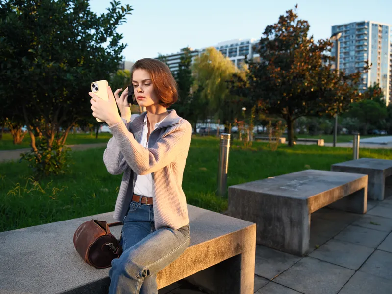 A focused young woman sits on a park bench, looking at her smartphone in a sunny urban setting. She appears thoughtful and connected, conveying a casual, modern lifestyle.