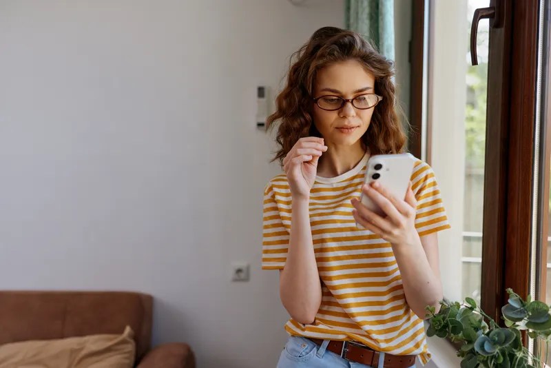 Casual woman indoors, looking at smartphone, striped tee, cozy home setting with warm natural light, relaxed posture, soft colors, greenery by window, candid lifestyle moment, contemporary interior