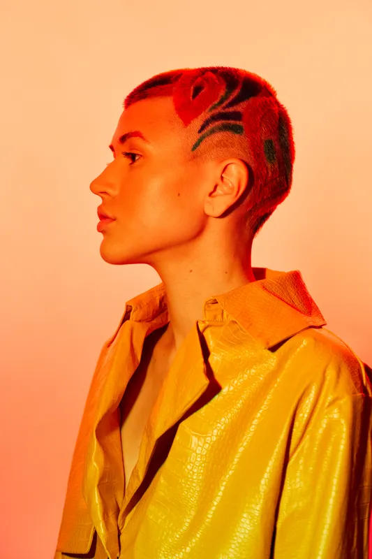 Young woman with short patterned hair and glowing makeup in bright yellow jacket, posing in warm orange lighting, stylish and bold look, creative portrait.