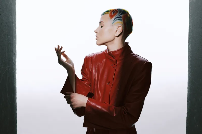 Young woman with artistic short hair and vibrant colors wearing a stylish red leather jacket poses confidently with a minimalistic white background, expressing creativity and modern fashion.