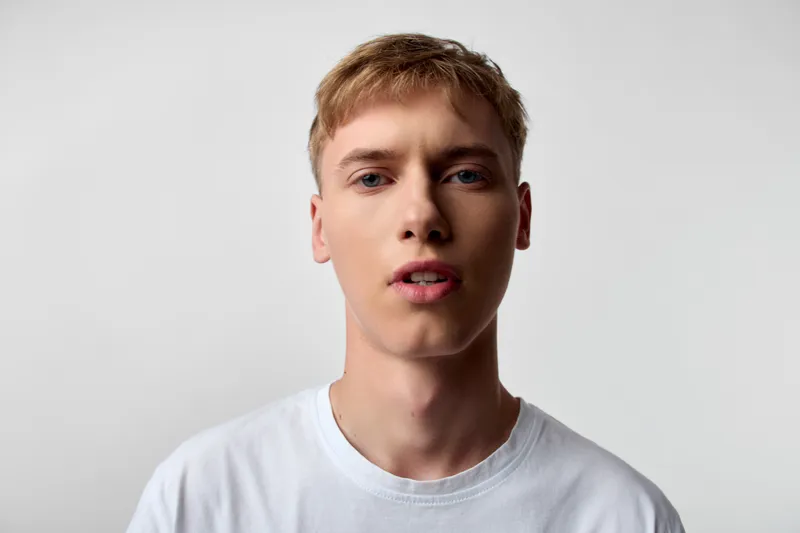 Young man with a neutral expression wearing a white t shirt against a plain studio backdrop, close up view highlighting facial features and skin tone for fashion, lifestyle or modeling use