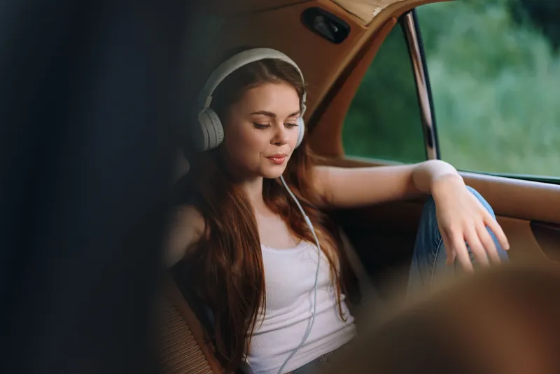 Young woman listening to music in a vintage car, wearing casual clothing, reflecting tranquility and joy with natural background.