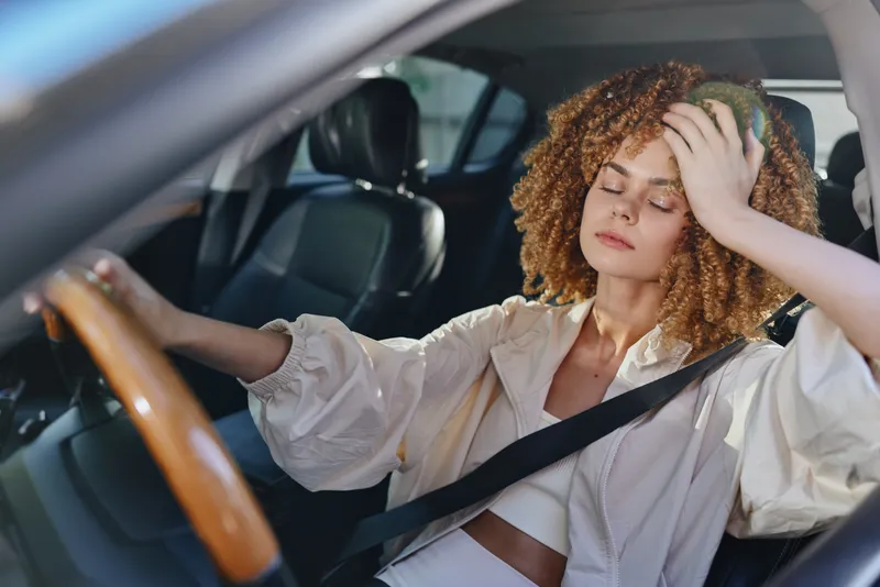 young woman driving car with curly hair looks stressed tired holding head inside modern vehicle interior in natural daylight