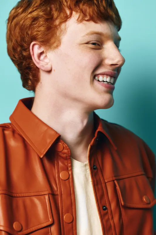 Young man with red hair smiling wearing brown leather jacket on blue background, happy portrait, side view of cheerful person in casual outfit.