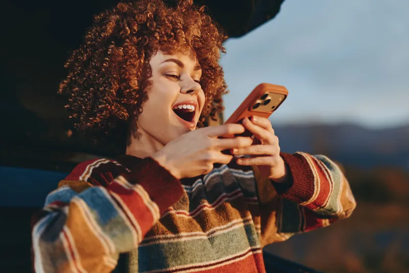 Happy woman lifestyle with curly hair laughing outdoors, holding smartphone in striped casual sweater, warm evening light, mountain landscape background, candid lifestyle moment