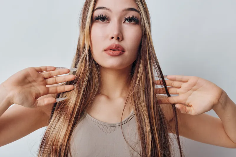 Young woman with long straight hair and neutral facial expression posing with raised hands in front of white background studio portrait for beauty concept.