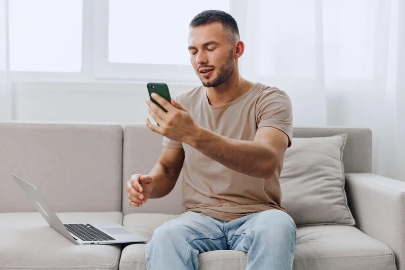 Engaging man using smartphone while sitting on a sofa, surrounded by natural light. The relaxed atmosphere reflects modern lifestyle trends of 2025-2026.