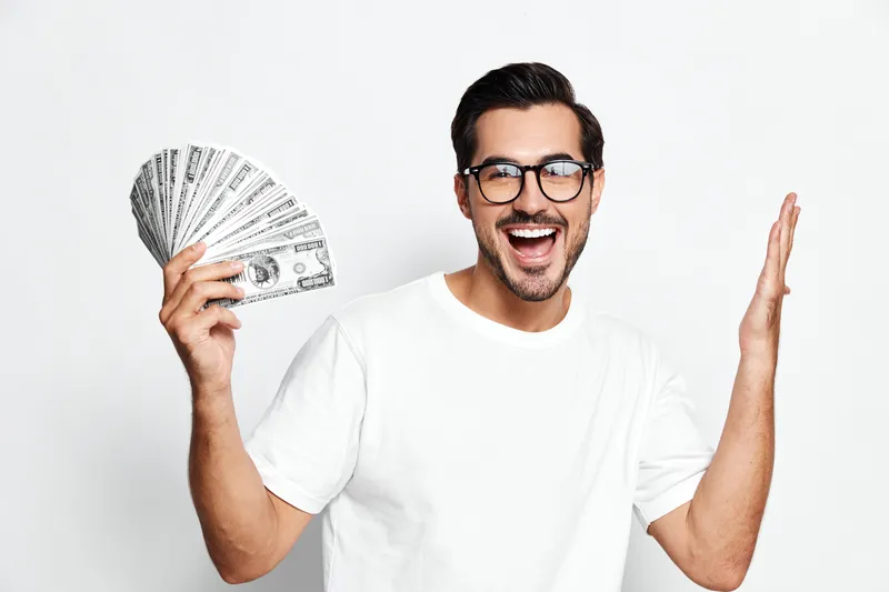 Happy cheerful young man in white t-shirt holding fan of dollar bills, looking excited and celebrating, isolated on plain solid background. Money success concept.
