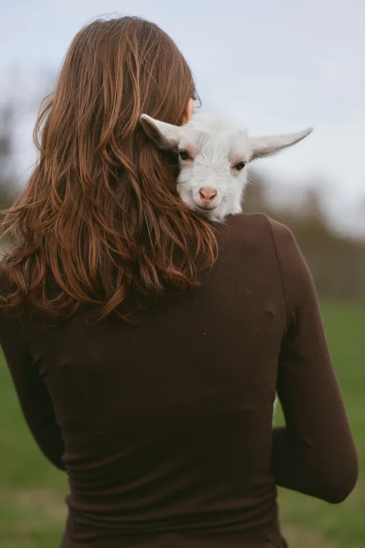 Young woman with long brown hair holds a small white goat on her shoulder outdoors, showing a gentle connection between human and animal in a natural lifestyle setting.