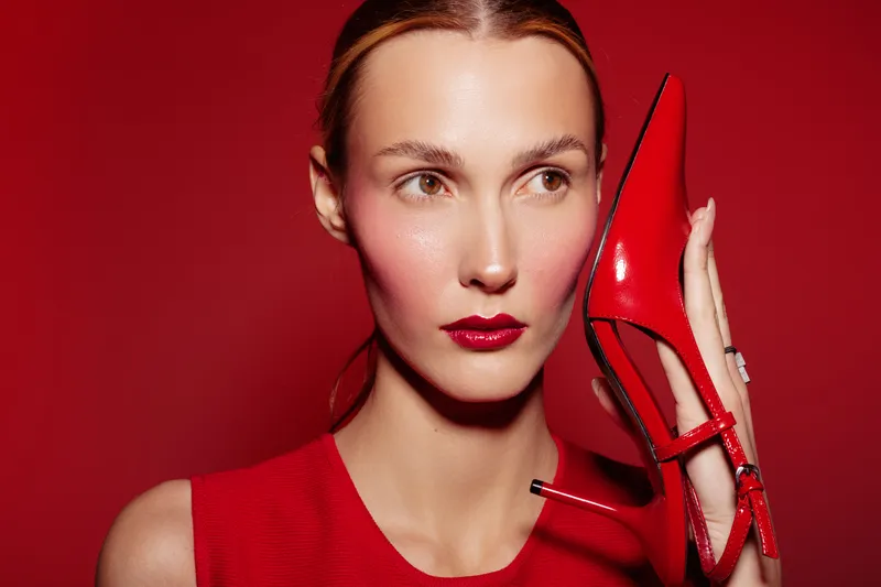 Elegant woman with red lipstick holds red high heel shoe near face against red background. Fashion concept with stylish makeup and matching outfit in vibrant studio lighting.