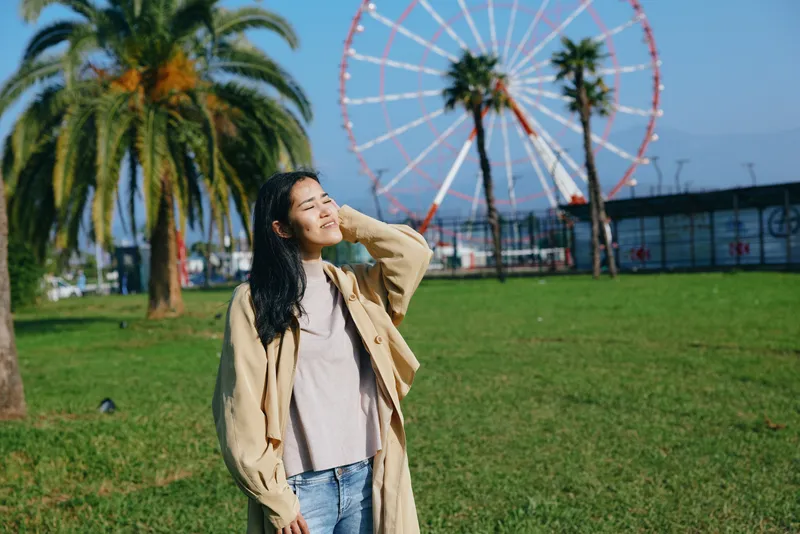Woman outdoors in a sunny park, wearing a beige coat and sweater, smiles while posing under a bright sky with a ferris wheel and palm trees in the background.