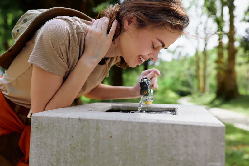Woman drinking from a concrete fountain in a park with backpack on shoulder, smile visible as she leans to hydrate outdoors; casual happy young traveler among trees in daylight refreshment.