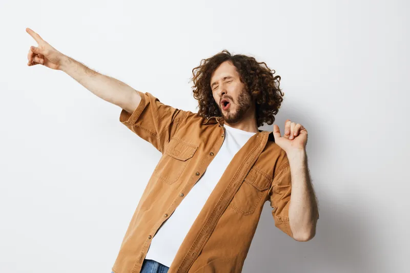 Young man expressing joy and enthusiasm while dancing against a minimalistic white background, dressed in a casual brown shirt and white t shirt, showcasing a carefree mood