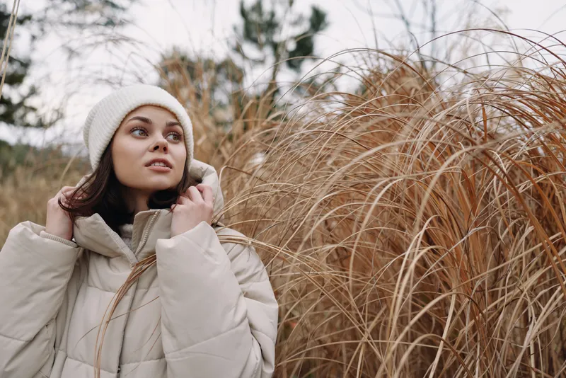 Woman in a warm beige coat and knit hat stands among tall dried grasses, captured in soft natural light, conveying calm solitude and seasonal mood in an outdoor portrait.