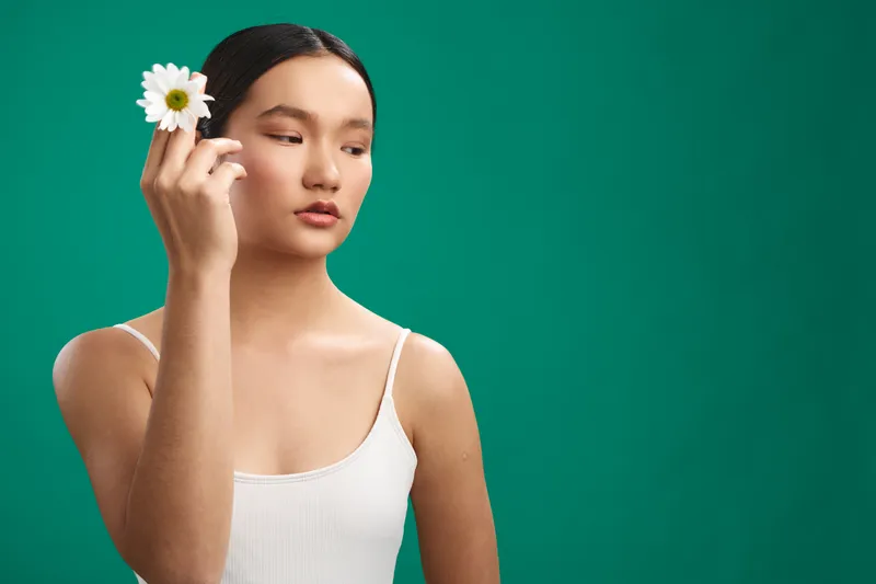 asian woman holds a flower gracefully in a studio portrait, calm expression, soft lighting, clean white top, vibrant green backdrop, simple composition conveys elegance, serenity, and contemporary