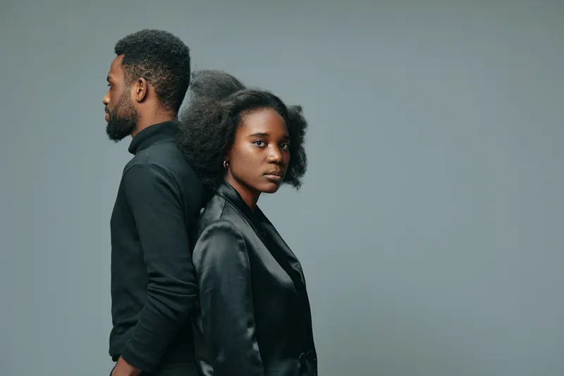 A young African couple standing back to back, expressing confidence and strength in a minimalist studio setting with a gray background